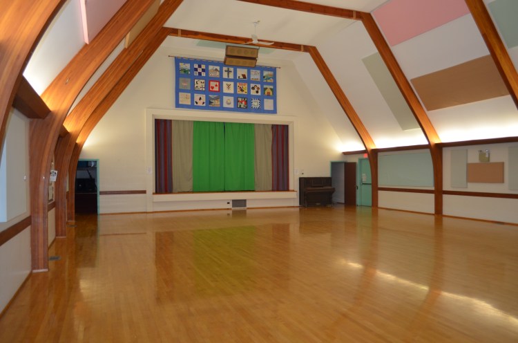 Bright community hall with wooden floors, vaulted ceiling and stage.